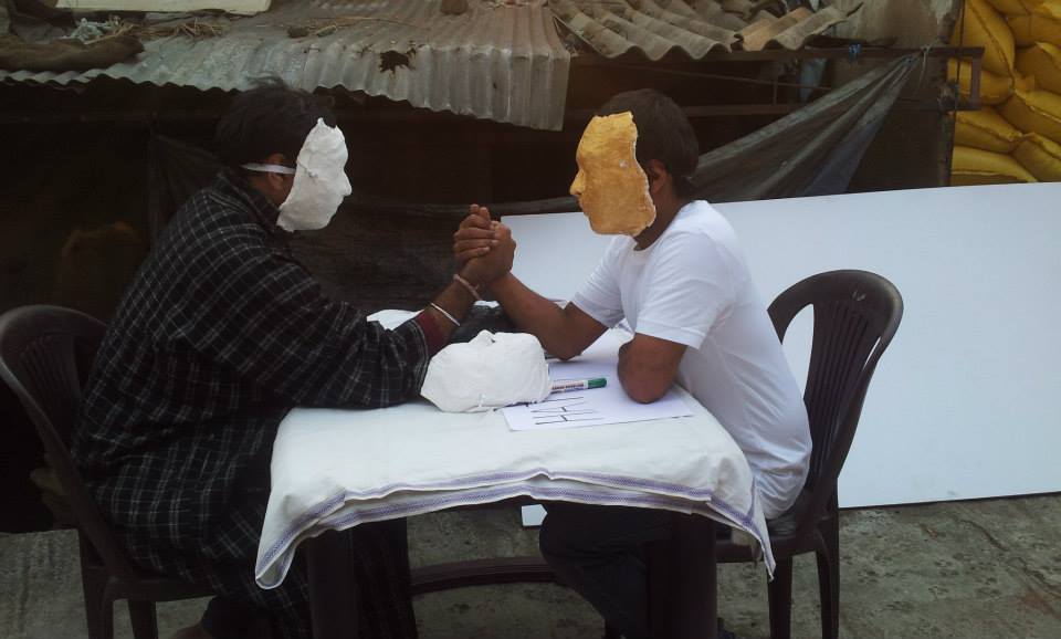 Jeetin Rangher wearing the mask of a 'Man' arm wrestles with 'Jealousy' (golden mask) Project 560: Art Adda at Sree Vinayaka Kalyana Mantapa, Bellary Road